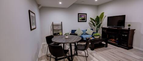 Una sala moderna con una mesa de comedor redonda, sillas negras, un sofá azul con cojines, un televisor sobre un mueble de madera y una estantería.