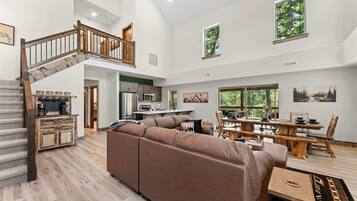 A spacious living area with a wooden staircase, a large sofa, a dining table, and a kitchen in the background.