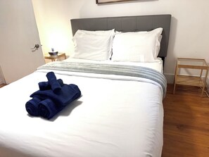 A neatly made bed with white linens and a blue towel, a grey headboard, a bedside table with a lamp, and a wooden nightstand. - Unit at the heart of Mississauga (Mississauga)