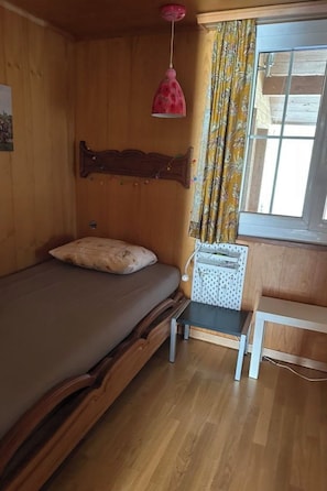 A wooden room with a bed, a small table, a hanging light fixture, and a window with curtains.