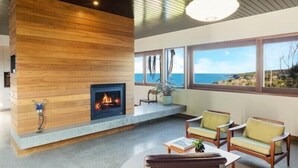 A modern living room with a fireplace, large windows, and a view of the ocean. - East Lane (Bega Valley)