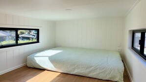 Een kamer met een schuine kap, houten vloer, een bed met een bedsprei met patroon, en twee ramen met uitzicht op de omgeving.