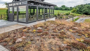 A modern glass structure with a metal roof, surrounded by a natural landscape with diverse vegetation.