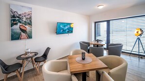A modern room with a wooden table, beige chairs, a TV, and a painting of a mountain lake.