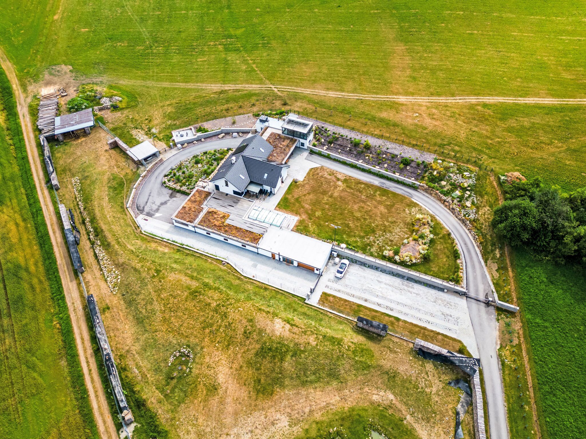 An aerial view of a modern building complex with a large parking area, a train track, and surrounding green fields.