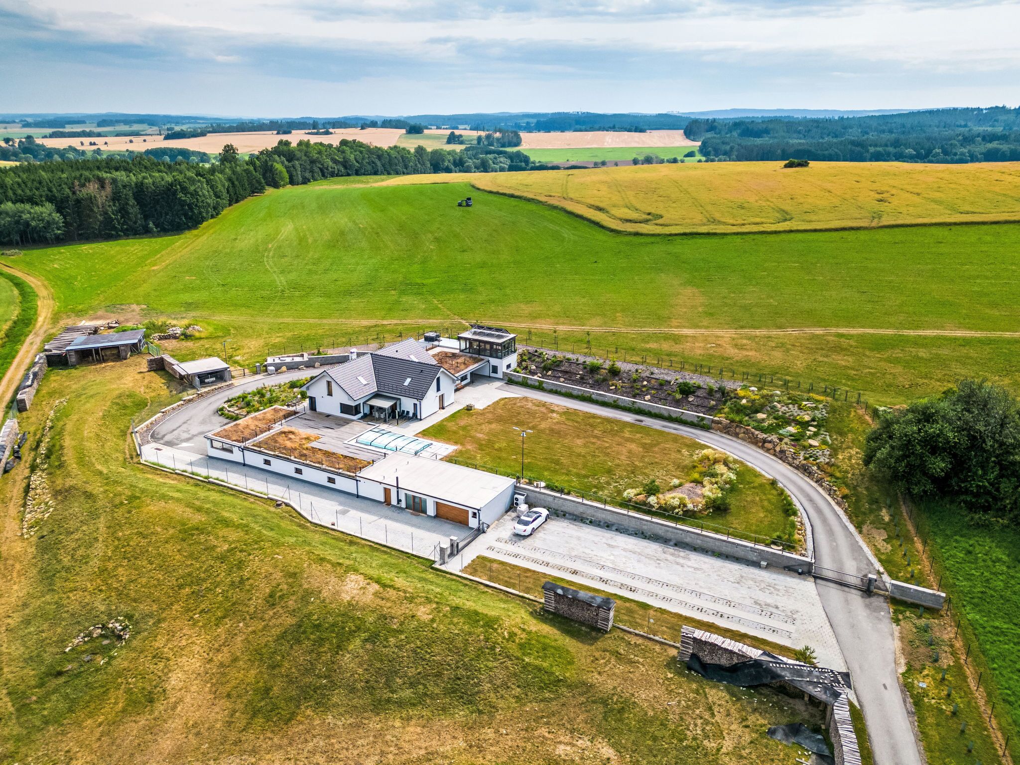 An aerial view of a modern building complex with a large parking area, surrounded by green fields and a curved road.