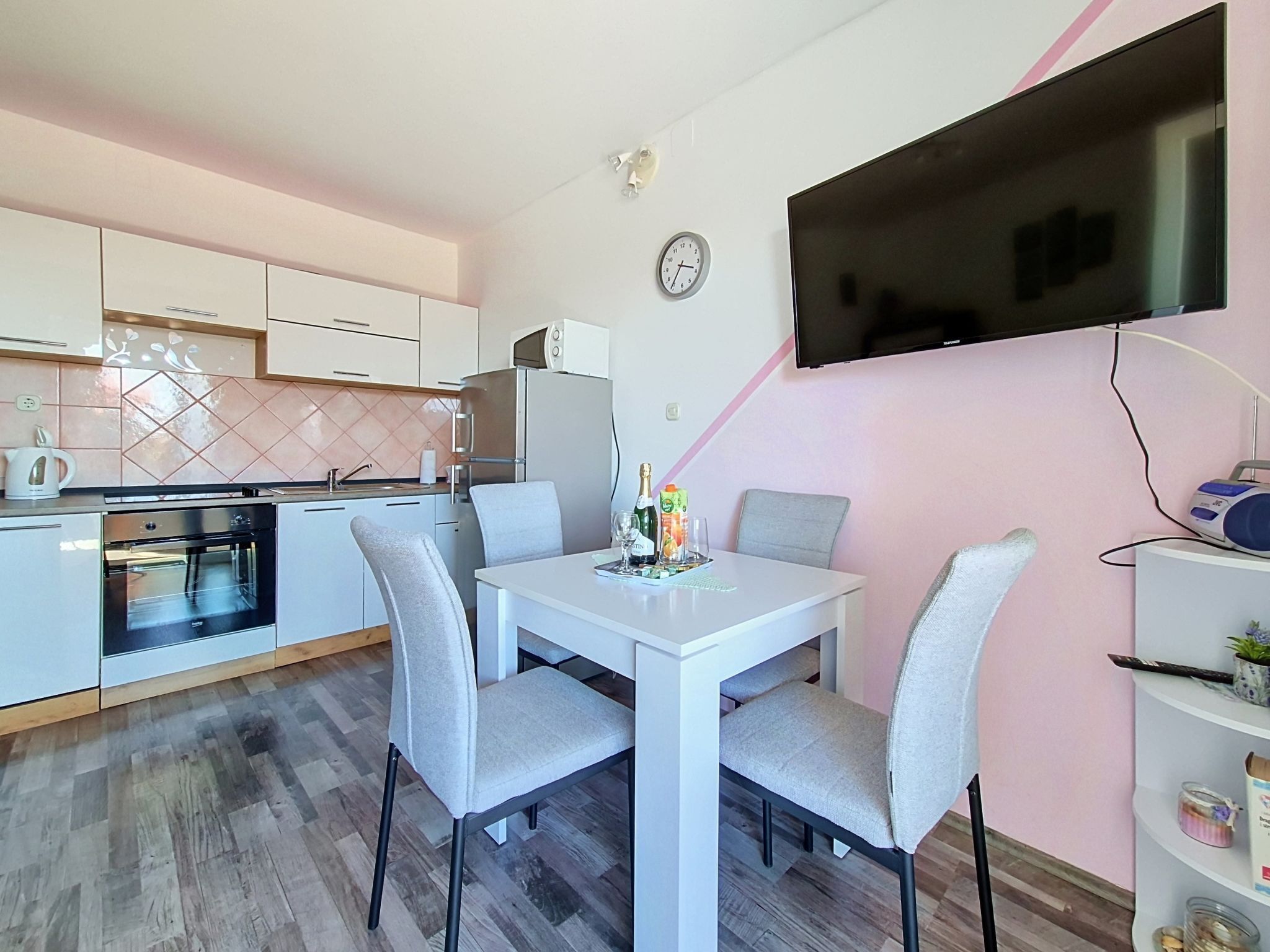 Uma cozinha moderna com armários brancos, forno embutido e um espaço de refeições com mesa branca e cadeiras cinzentas.
