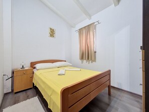 Un dormitori amb un llit de fusta, una tauleta de nit, una làmpada penjada a la paret, una finestra amb cortines i una petita peça decorativa a la paret.