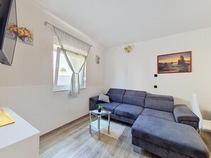 Un saló amb un sofà seccional gris, una taula petita de vidre amb una planta, una finestra amb cortines estriades i un quadre del capvespre a la paret.