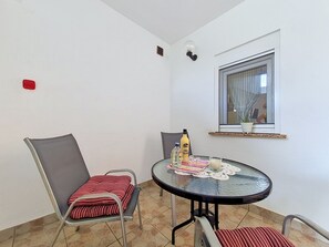 A small dining area with a round table, two chairs, a window with a curtain, and a potted plant on the windowsill. - Apartment Vigi by Interhome (Pag/Povljana)