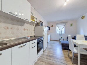 A modern kitchen with white cabinets, a built-in oven, and a microwave. There is a dining area with a white table and chairs, and a sofa in the background. - Apartment Vigi by Interhome (Pag/Povljana)