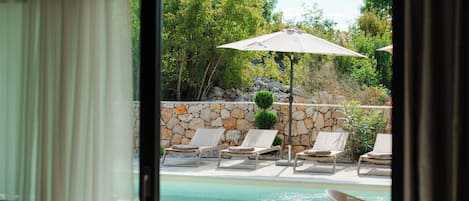 A view of a swimming pool with lounge chairs and umbrellas, surrounded by a stone wall and greenery.