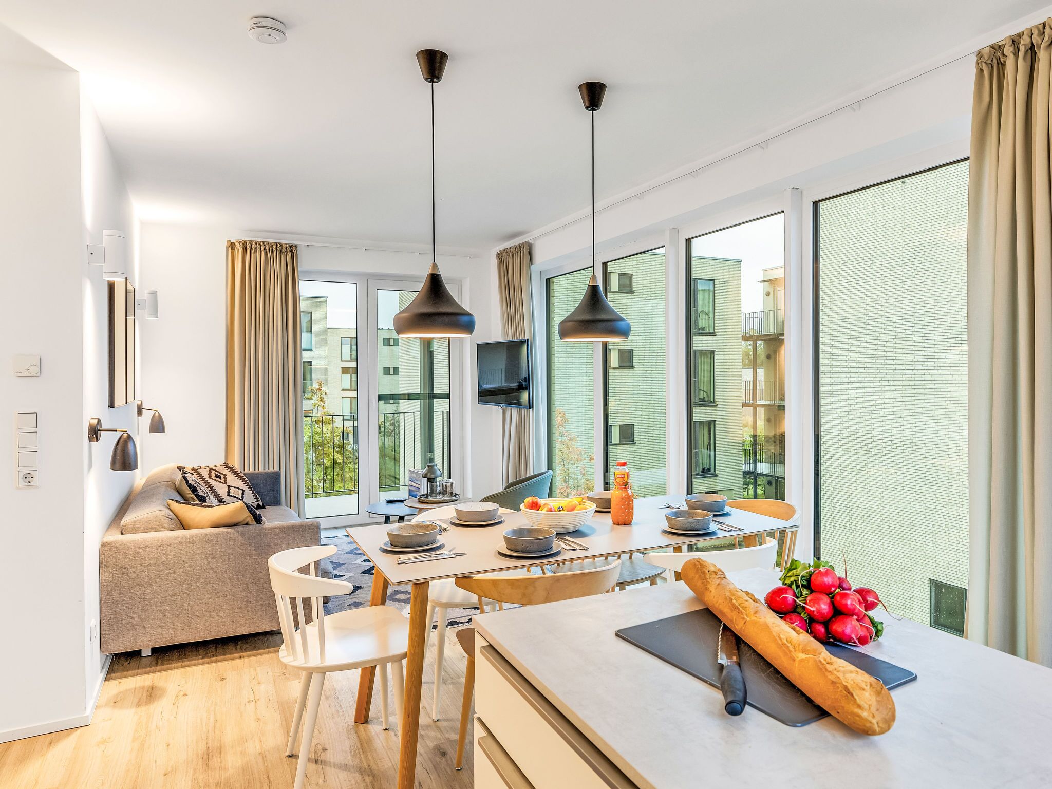 Une cuisine moderne avec une table à manger, des chaises et un canapé. De grandes fenêtres sont présentes, ornées de rideaux, et un luminaire est fixé au plafond. On peut admirer la vue sur les bâtiments environnants.