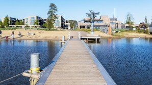 En træbro der strækker sig ud i roligt vand, med moderne bygninger og en sandstrand i baggrunden.