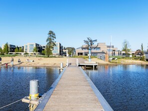 Eine Holzstege, der sich in ruhiges Wasser erstreckt, mit modernen Gebäuden und einem Sandstrand im Hintergrund.