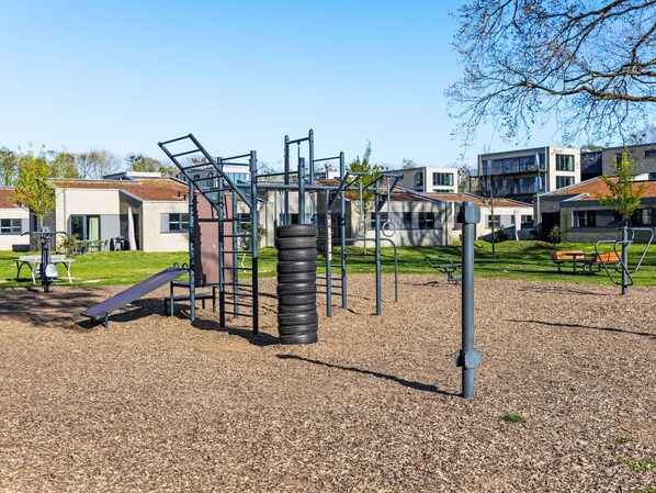 Um parquinho com estruturas de escalada e um escorregador, cercado por uma área de brita e edifícios residenciais ao fundo.