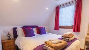 A bedroom with a bed, pillows, a nightstand, a window with curtains, and a shelf with towels. - Invergarry Combi by Interhome (Invergarry)