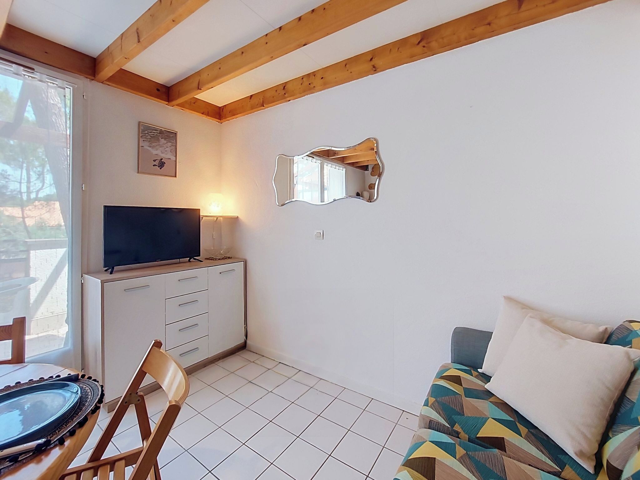 Ein Zimmer mit Fernseher auf einer Kommode, einem Spiegel an der Wand, einem gemusterten Sofa und einem Sessel.