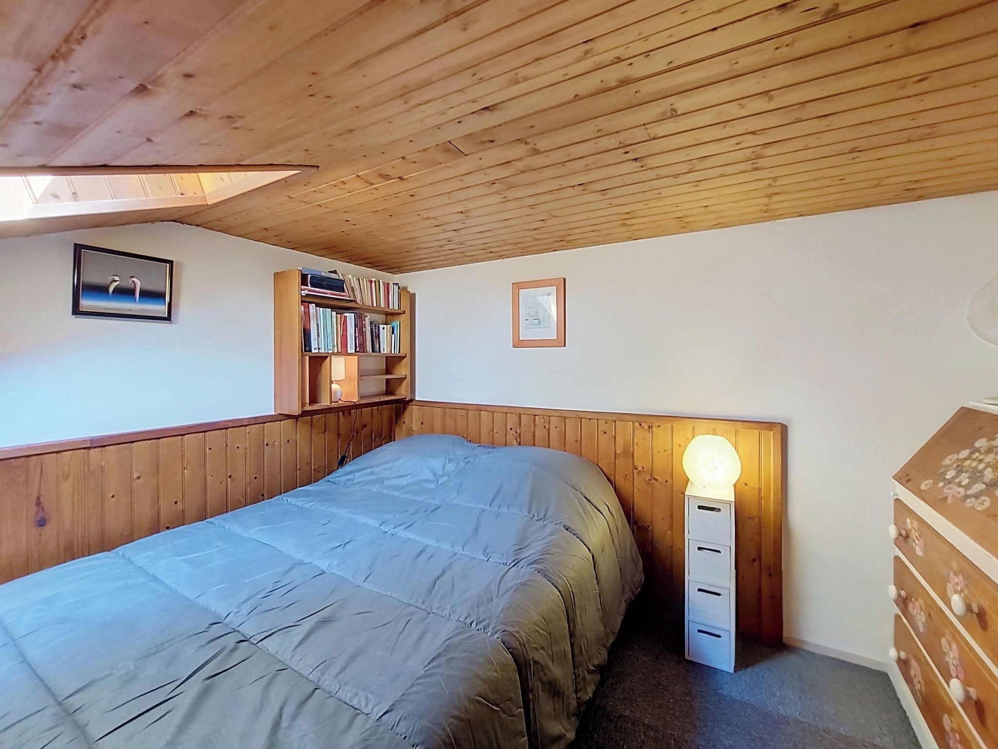 Un dormitorio con techo de madera, una cama con edredón gris, una lámpara de noche, una estantería y un aparador.