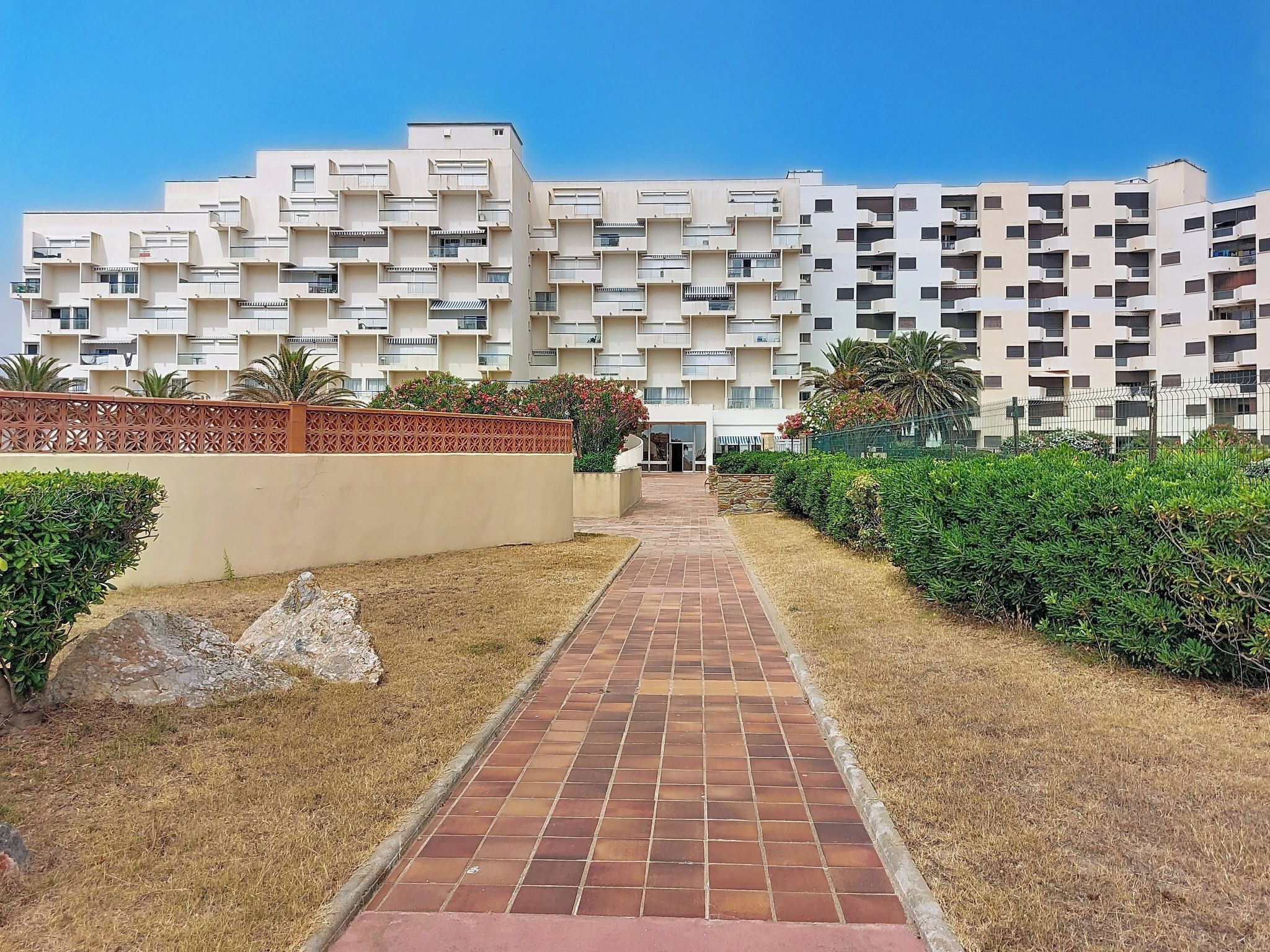 Un edifici d'apartaments modern amb entrada central, envoltat de jardineria ben cuidada i un passeig pavimentat.