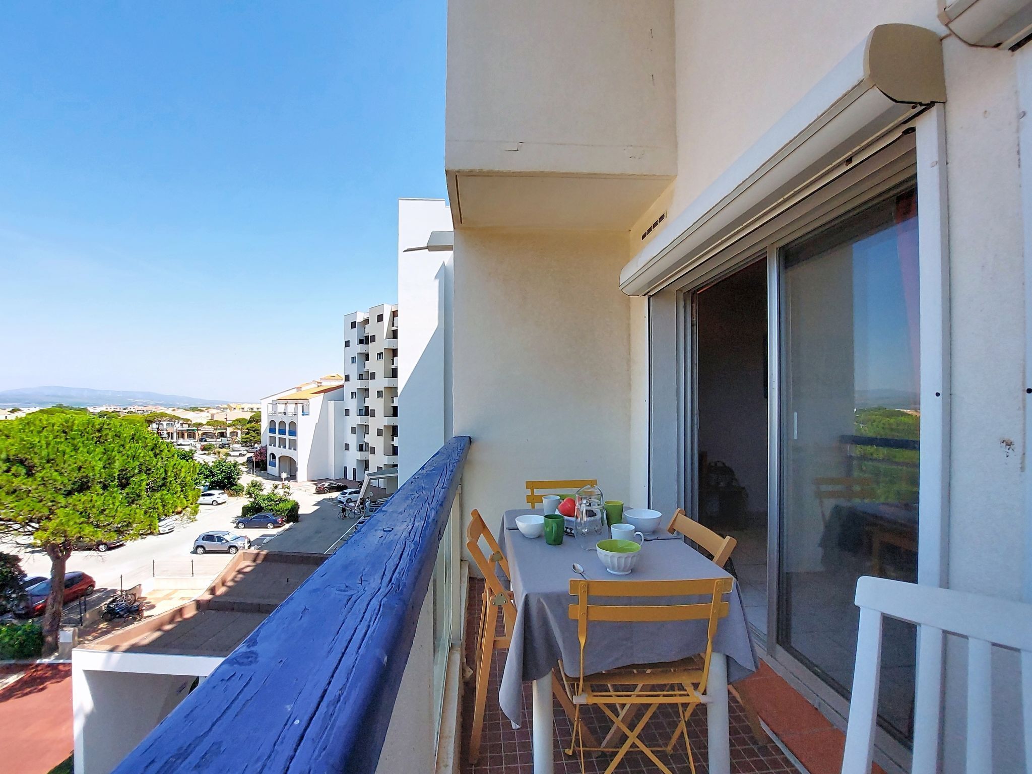 A balcony with a table set for dining, overlooking a street with parked cars and buildings.