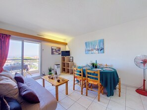 A furnished living room with a sofa, a dining table set for four, a TV, and a fan. - Lydia Playa by Interhome (Le Barcarès)