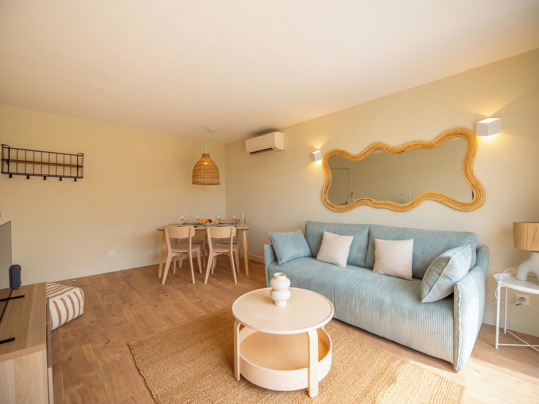 A modern living room with a sofa, a small round table, a dining table with chairs, a hanging light fixture, and a wall-mounted mirror.
