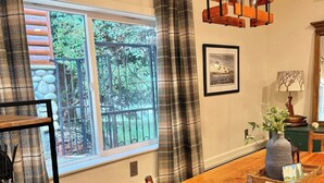 A dining area with a wooden table, chairs, a hanging light fixture, a window with curtains, and a view of greenery outside. - Lux Lodge Downtown w/ AC, Bonus Cottage, Courtyard (McCall)