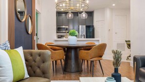 Un comedor moderno con una mesa redonda, cuatro sillas, una araña y una cocina al fondo.