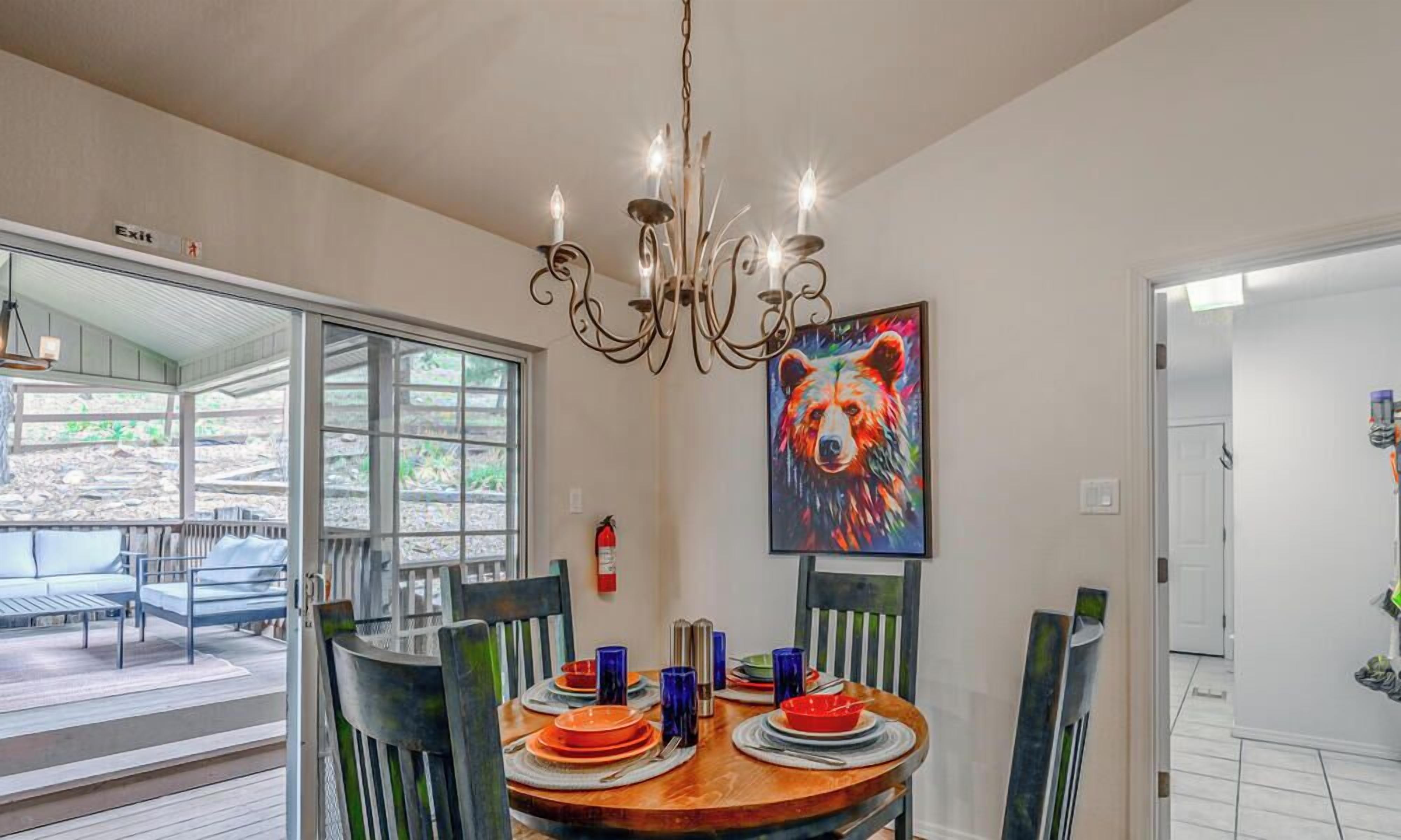 A dining area with a round table, chairs, and a chandelier. There is a painting on the wall and a fire extinguisher nearby.