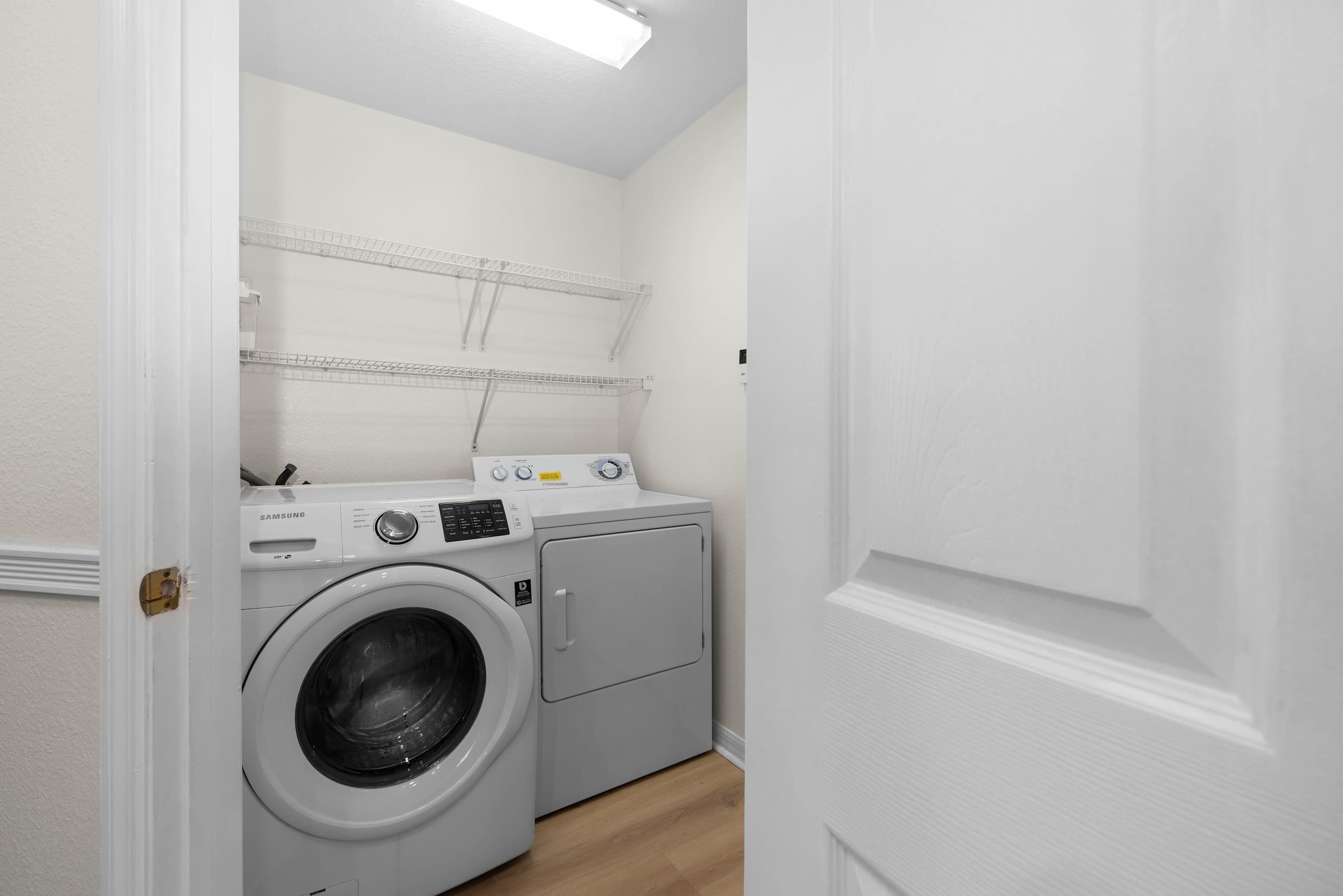 Une buanderie équipée d’un lave-linge et d’un sèche-linge à chargement frontal, avec des étagères au-dessus et une porte donnant sur une autre pièce.