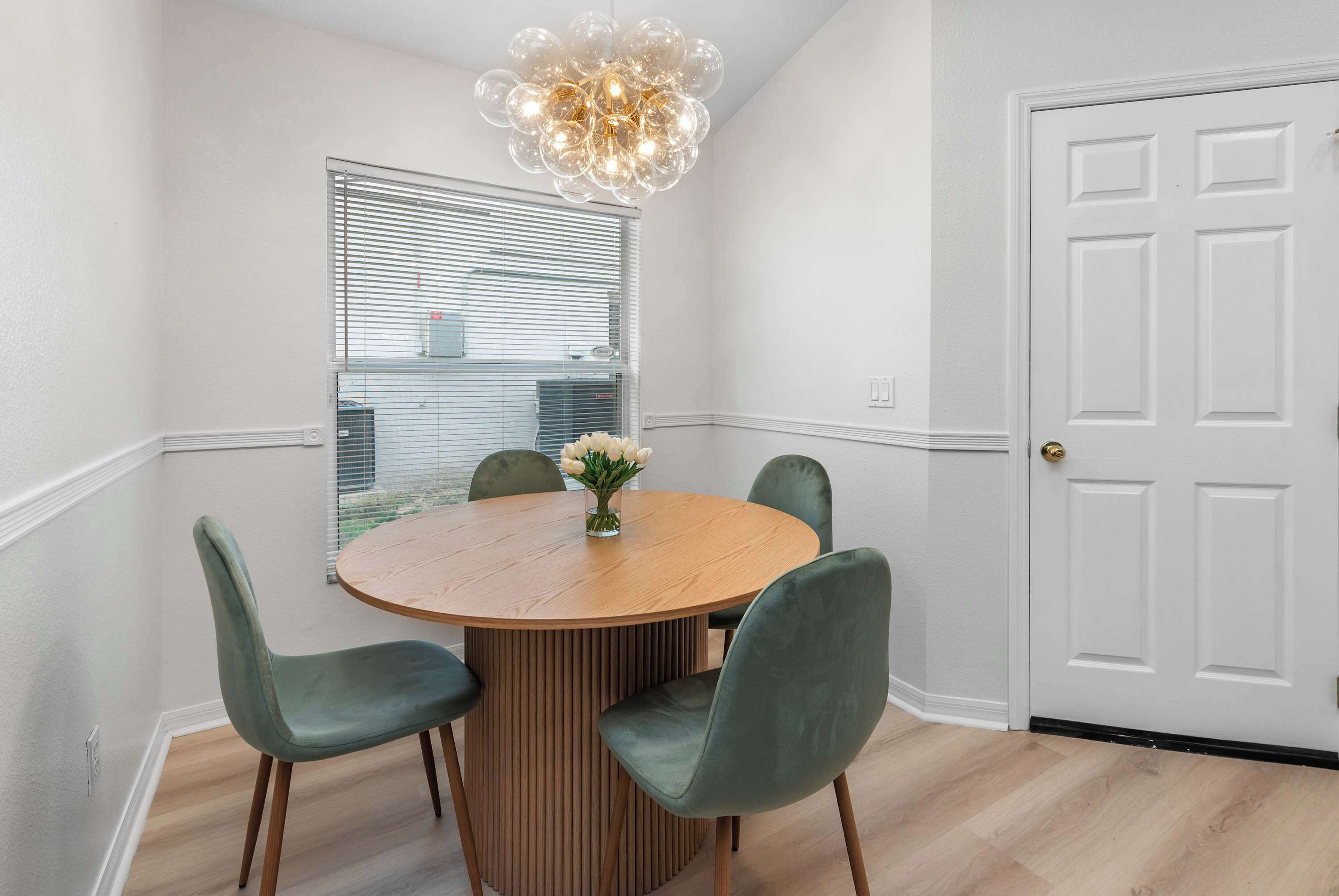 Een eetgedeelte met een ronde houten tafel, vier stoelen, een vaas met bloemen, een raam met zonwering en een witte deur.