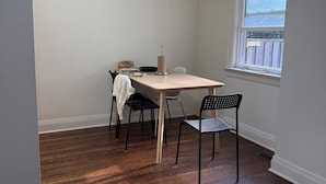 Una habitación con suelo de madera, una mesa con sillas, una ventana con persianas y una lámpara en el techo.