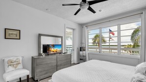 Une chambre à coucher comprenant un lit, une télévision, une commode, une chaise et une fenêtre donnant sur la plage et les palmiers.
