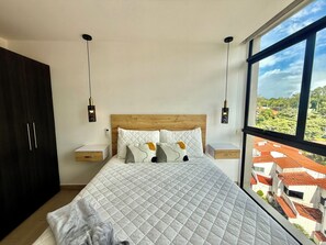 Ein Schlafzimmer mit einem großen Bett, zwei Nachttischen, einem Kleiderschrank und Blick auf Gebäude durch ein Fenster.