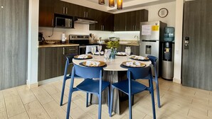 Een moderne keuken met donkere houten kastjes, een roestvrijstalen koelkast, een magnetron en een gasfornuis. Er staat een eettafel met blauwe stoelen en erboven een kroonluchter.