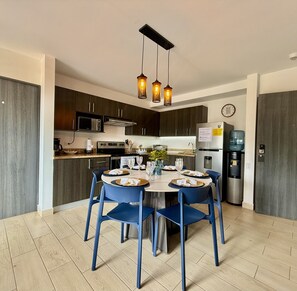 Una cocina moderna con armarios de madera oscura, un refrigerador de acero inoxidable, un microondas y una estufa a gas. Hay un comedor con sillas azules y una lámpara de araña colgante.