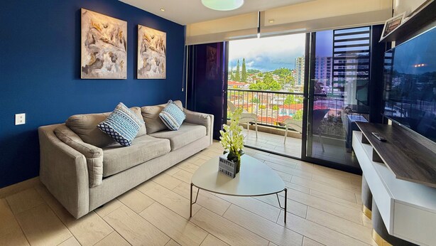Een moderne woonkamer met een bank, een salontafel en een televisie. Twee schilderijen hangen aan de muur, en er is een balkon met uitzicht op de stad.