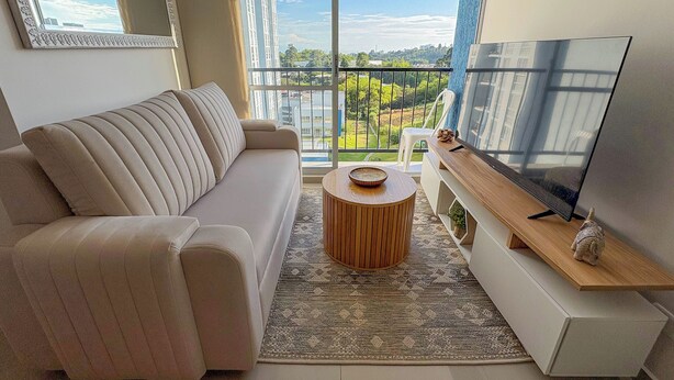 Un salon moderne avec un canapé, un téléviseur, une petite table ronde et un balcon offrant une vue sur les bâtiments et la verdure.