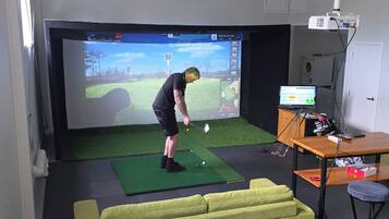 Una habitación con una pantalla grande mostrando un partido de golf, un sofá verde, un escritorio con una computadora y un carrito de golf.