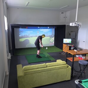 A room with a large screen displaying a golf game, a green couch, a desk with a computer, and a golf cart. - Stay n Play golf sim and game room adventures in the heart of Pukekohe (Pukekohe)