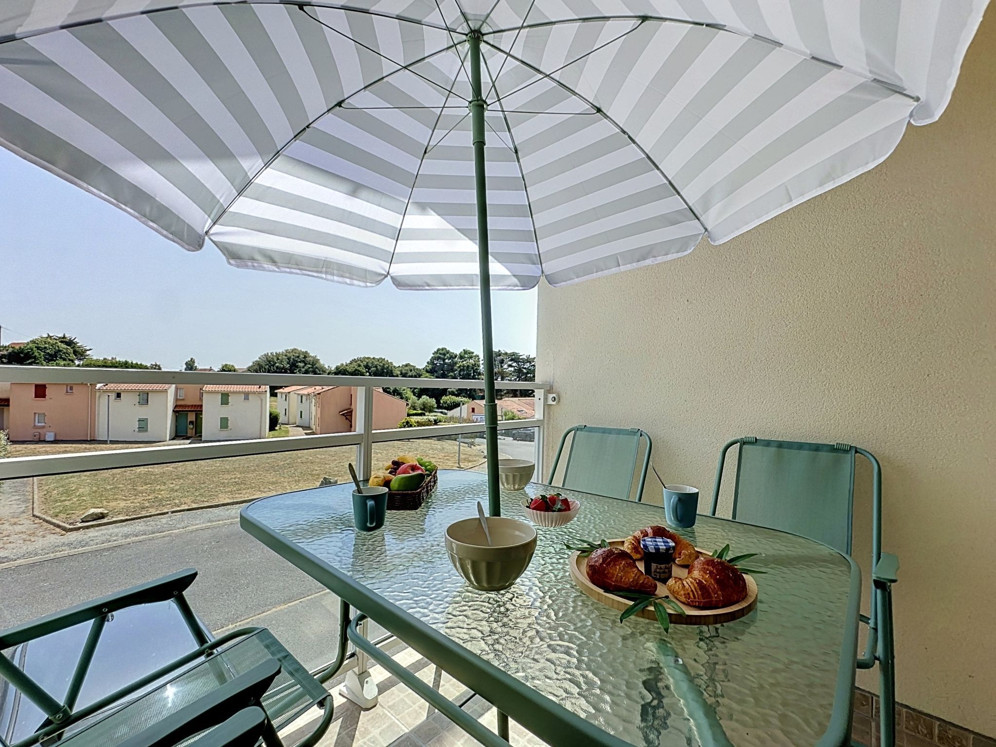 Een glazen tafel met stoelen, een strepen paraplu en een schaal fruit op een balkon.