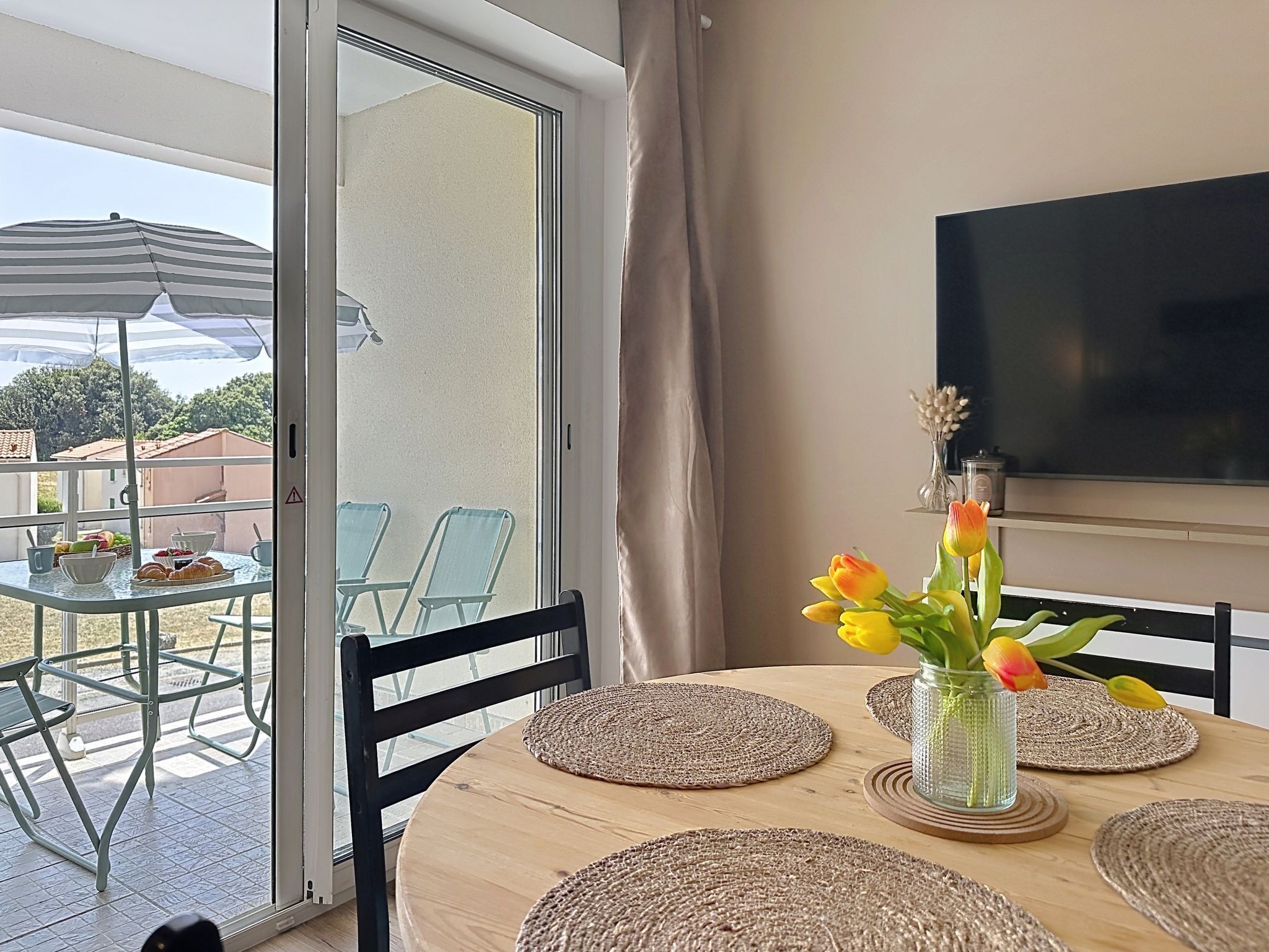 Un espace repas comprenant une table, des chaises, un téléviseur et une vue sur l’extérieur, visible à travers une porte vitrée.