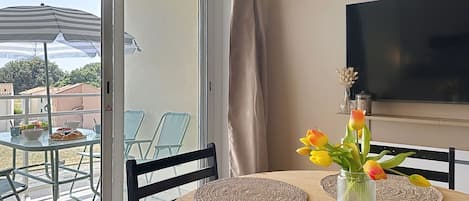 Un espace repas comprenant une table, des chaises, un téléviseur et une vue sur l’extérieur, visible à travers une porte vitrée.