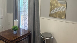 A room with a wooden table, a trash can, a wall-mounted radiator, and a window with patterned curtains.