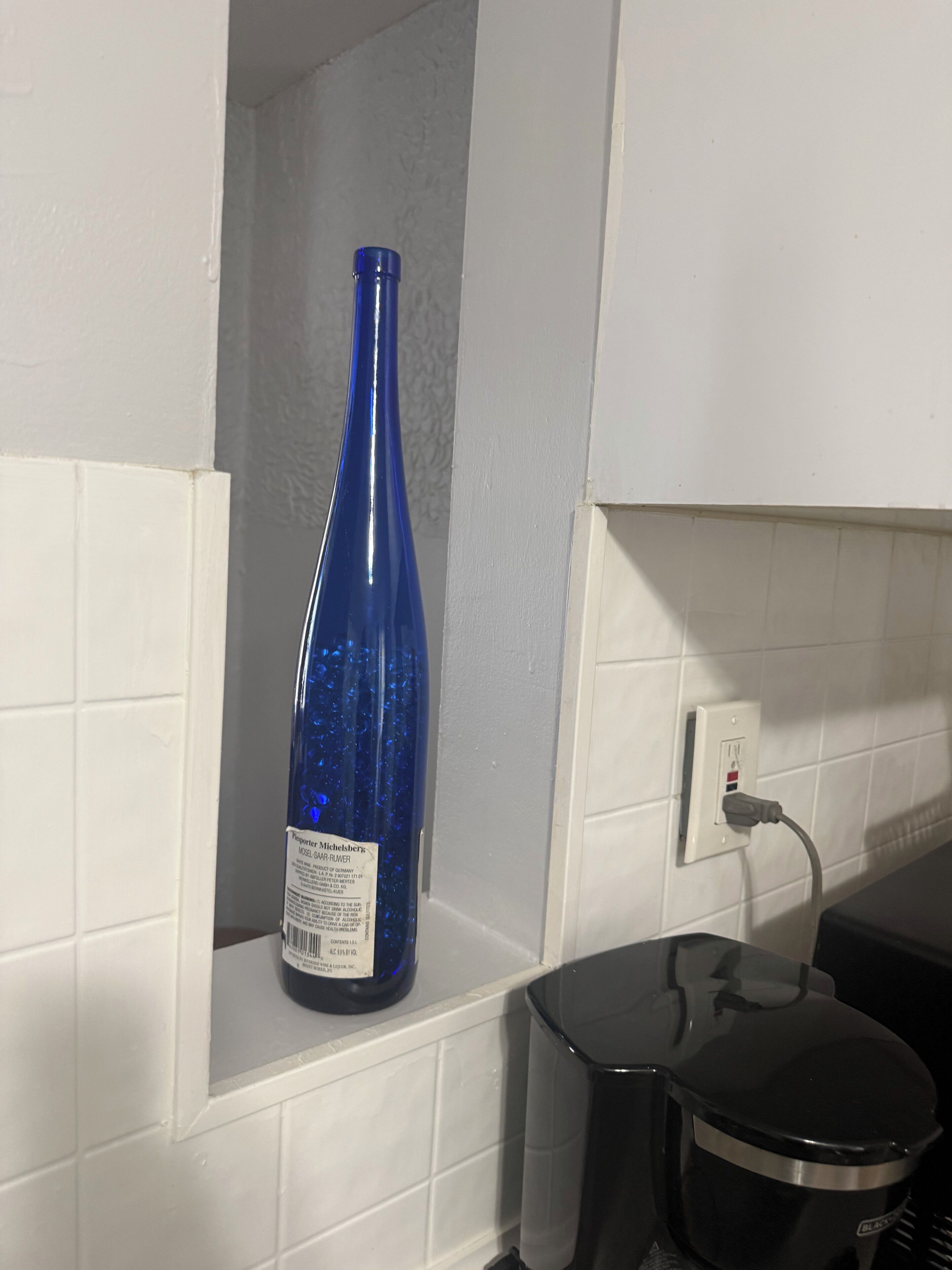 A blue bottle on a shelf, a black appliance, and a white tiled wall with a power outlet.