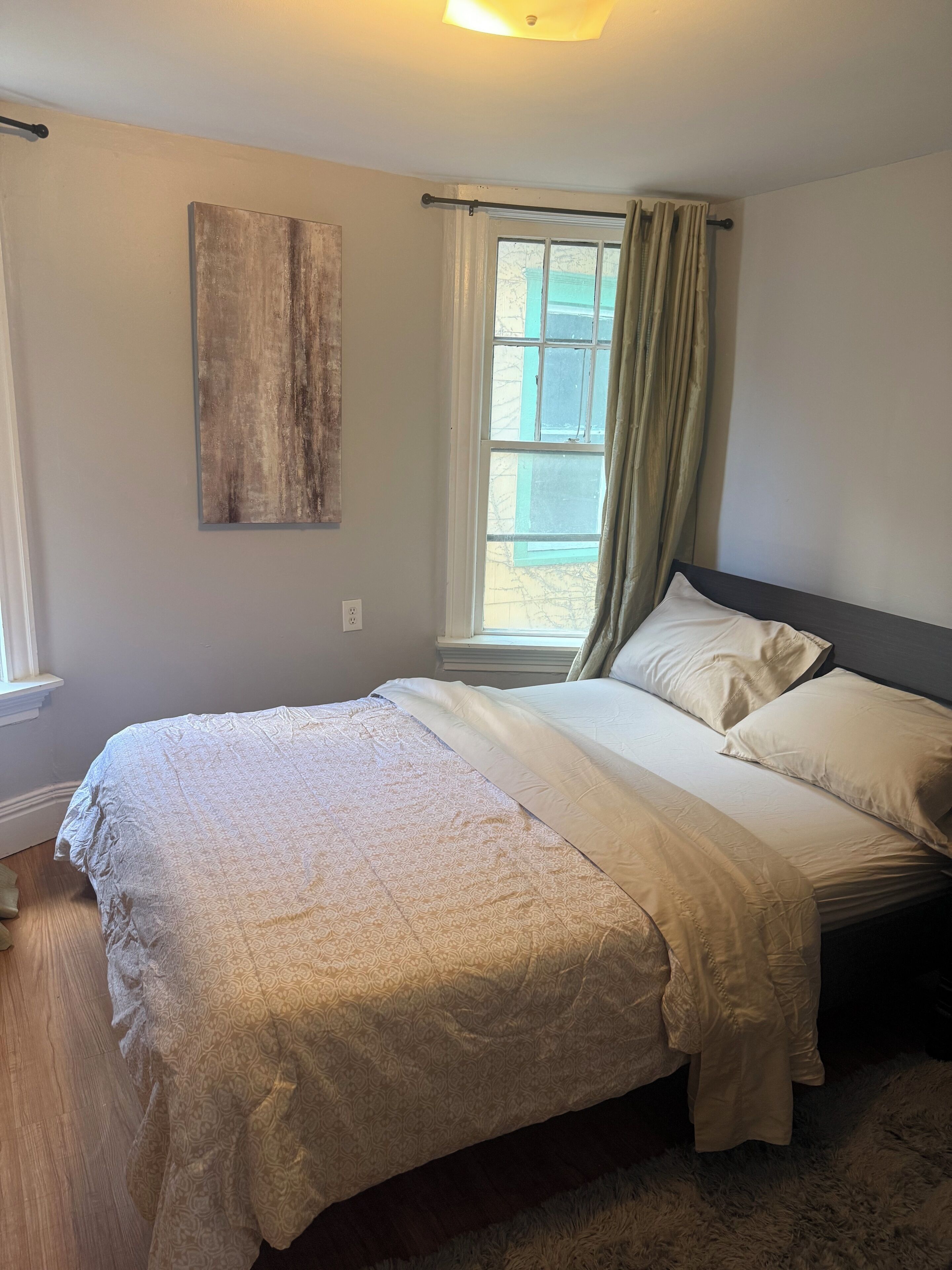Quarto com cama, janela com cortinas, uma obra de arte na parede e luminária.