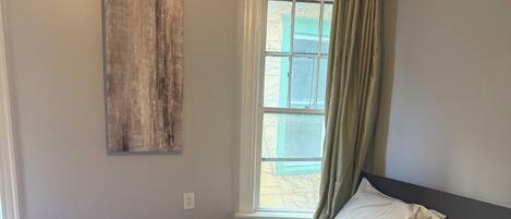 Quarto com cama, janela com cortinas, uma obra de arte na parede e luminária.