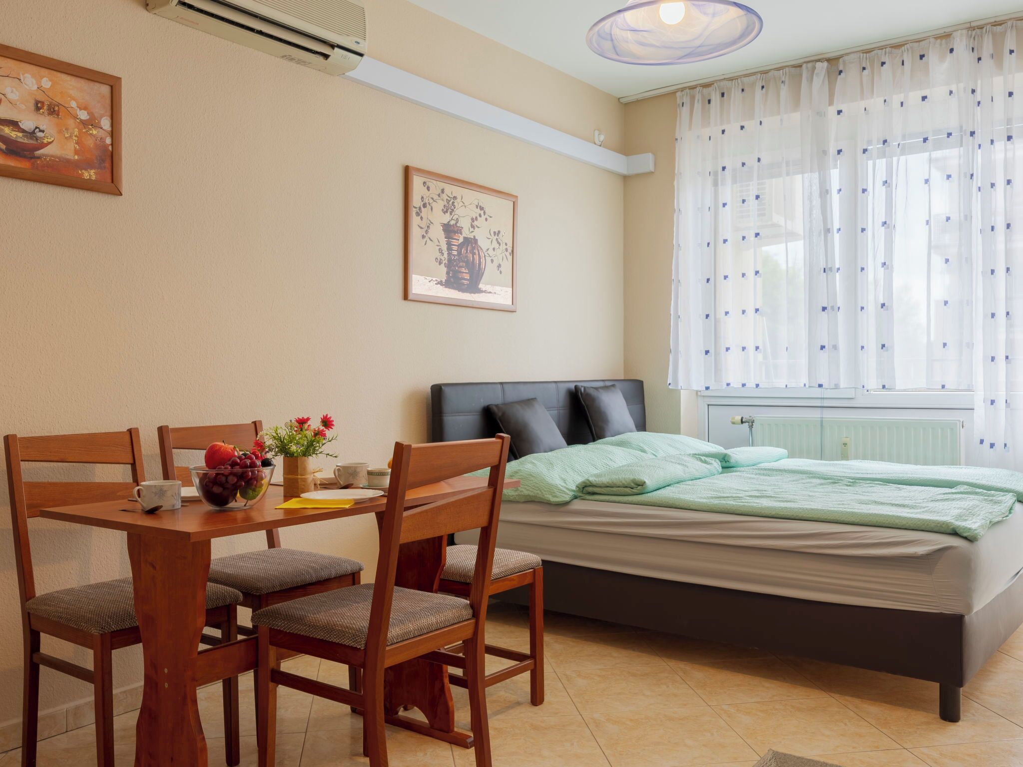 Een slaapkamer met een bed, een eettafel met stoelen, een raam met gordijnen en een radiator.