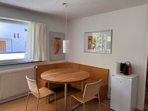 A compact room with a round wooden table, two wicker chairs, a white radiator, a small white fridge, and a window with curtains. - Solaria 31 Ferienhaus, Dusche oder Bad, WC 5 1/2- Ferienhaus by Interhome (Davos)